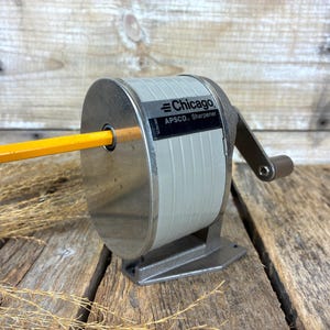 May include: A vintage Chicago APSCO pencil sharpener with a light gray cylindrical body, a metallic silver front plate, and a manual hand crank. A yellow pencil is inserted into the front opening. The device features a dark base and a label.