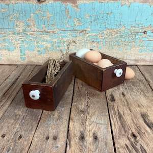 May include: Two rectangular dark brown wooden drawers with white ceramic knobs. One drawer contains dried plant stems, while the other holds several light brown and white eggs. The drawers are constructed from wood with visible grain and dark finish.