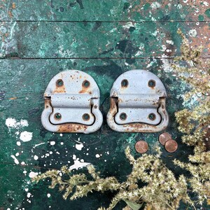 May include: Two matching metal chest handles with a weathered, light gray painted finish and visible rust patches. Each handle features a rounded top section with two screw holes and a hinged, U-shaped pull mechanism with two additional screw holes.