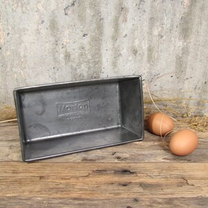 May include: A rectangular metal bread pan with the word "Morton" embossed on the side. Two brown eggs and a few strands of dried grass are next to the pan. The pan is on a wooden surface.