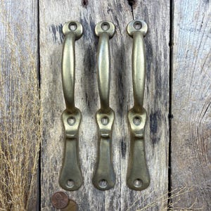 May include: Three matching brass door handles with a curved grip and an elongated base plate. Each handle features three circular screw holes for mounting, with one hole at the top and two at the bottom of the plate. The metal has a muted gold finish.
