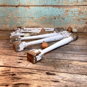 May include: A collection of white, distressed wooden spindles with square bases, arranged on a weathered wooden surface. The spindles have peeling paint, revealing the wood underneath. Several brown and green eggs are scattered around the spindles, suggesting a rustic, farmhouse theme.