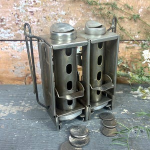 May include: A vintage dual-tube metal coin dispenser with a silver-toned finish. The device features two vertical cylindrical chambers with side slots and hinged top caps. Several round metal coins are resting at the base of the dispenser.