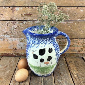May include: A ceramic pitcher featuring a blue and white sponge-painted finish with a hand-painted black and white cow illustration on the front. The pitcher contains a sprig of dried herbs and is positioned next to two brown chicken eggs.