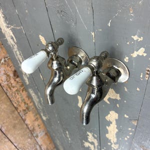 May include: A pair of wall-mounted faucets with polished silver-tone metal bodies and white ceramic handles. Each handle features black text, with one marked HOT and the other marked COLD, and both include circular metal base plates for installation.