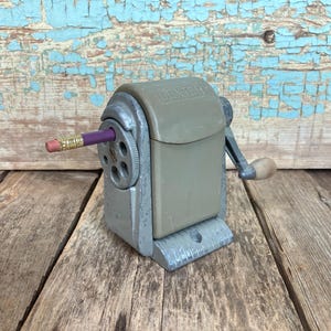 May include: A vintage metal pencil sharpener with a crank handle. The sharpener is gray and has the word "Dexter" printed on the side. A purple pencil is sticking out of the sharpener.