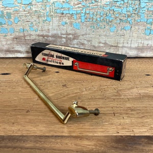 May include: A vintage brass furniture pull handle with a rectangular design, next to a black and red box labeled "Furniture Hardware". The box has the brand name "Faultless". The handle is on a wooden surface.