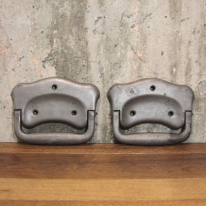 May include: Two vintage metal drawer pulls with a gray finish. Each pull has a curved handle and a decorative top with three holes for mounting. The pulls are displayed on a wooden surface against a textured concrete wall.