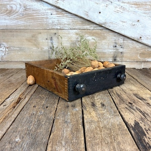 May include: A rustic wooden drawer with a black front and two dark knobs. The drawer is filled with walnuts and dried foliage. The drawer sits on a weathered wooden surface, suggesting a vintage or farmhouse aesthetic.