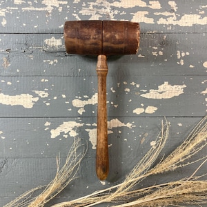 May include: A vintage wooden mallet with a cylindrical head and a long, slender handle. The mallet is made of dark brown wood, showing signs of wear and age. The background is a weathered, gray-blue wooden surface.