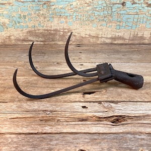 May include: An antique, dark metal hay hook with three curved tines and a handle. The tool is set against a weathered wooden surface with a distressed, light blue painted background. The hook appears to be well-worn.