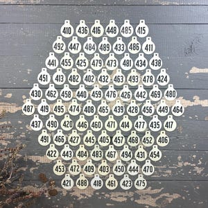May include: A collection of circular off-white tags arranged in a large hexagonal pattern. Each tag features a small hole at the top and is printed with a unique three-digit number in a bold black sans-serif font, ranging from 402 to 495.