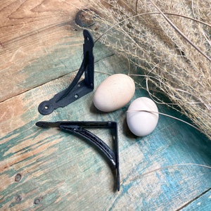 May include: Two black metal shelf brackets with a curved design. The brackets are on a wooden surface with two white eggs.