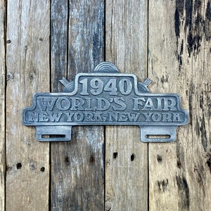 May include: A weathered metal sign from the 1940 World's Fair in New York, New York. The sign is rectangular with the text "1940 WORLD'S FAIR NEW YORK, NEW YORK" in raised lettering. The sign is mounted on a rustic wooden background.