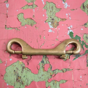May include: A brass swivel snap hook with two loops. The hook is on a pink and green background with peeling paint.