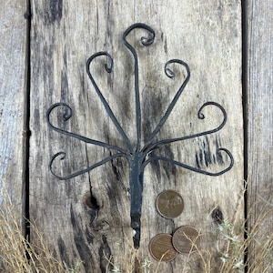 May include: A handcrafted, dark metal decorative piece with a central stem and stylized, curved arms. The piece is placed on a weathered wooden surface, accompanied by three pennies. The design evokes a natural, organic aesthetic.