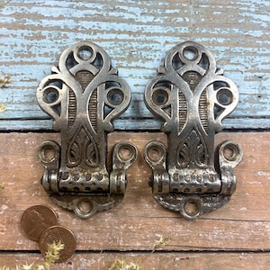 May include: Two antique silver-toned metal hinges with ornate, symmetrical designs. Each hinge has a decorative top and a hinged base. Two copper pennies are near the hinges. The hinges are on a weathered wooden surface.