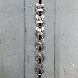 May include: A vertical arrangement of six silver-toned ring pulls, each with a circular handle and a decorative backplate. The pulls are mounted on a weathered, light gray wooden surface, creating a vintage aesthetic.