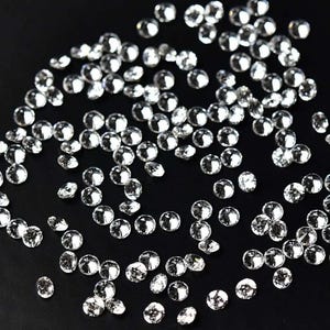 May include: A scattering of small, clear, round gemstones with multiple facets, reflecting light. The gemstones are scattered across a black surface, creating a sparkling effect. The gems are likely used for jewelry or craft projects.