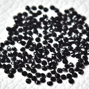 May include: A large pile of black round gemstones, likely diamonds, with a faceted cut. The gemstones are scattered on a white surface.