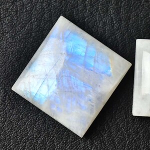 May include: Two white, square-cut moonstone gemstones with iridescent blue and white flashes, next to a U.S. quarter. One stone is a diamond shape. The gemstones are on a black surface.