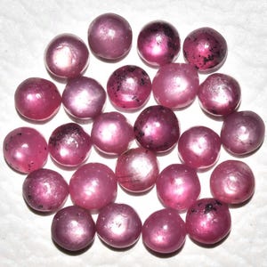 Op de afbeelding: Een verzameling ronde, roze edelstenen met een enigszins doorschijnend uiterlijk. Sommige stenen hebben donkere insluitsels. De edelstenen zijn in een cirkelvormig patroon gerangschikt, waarbij hun gepolijste oppervlakken en natuurlijke variaties worden getoond.