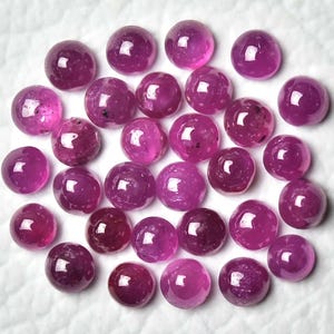 May include: A collection of round, polished gemstones in varying shades of pink and purple. The gemstones are scattered across a white surface, showcasing their smooth, reflective surfaces. The gems appear to be cabochons.