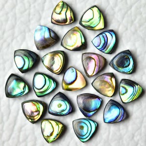 以下が含まれることがあります： アワビの貝殻で作られた、虹色の三角形の宝石カボションのコレクション。磨かれた貝殻は、青、緑、紫など、さまざまな色合いと渦巻き模様を示しています。カボションは白い表面に配置されています。