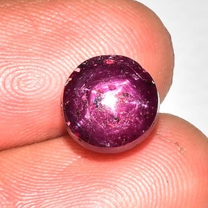 May include: A round, polished gemstone with a deep magenta hue. The gem has a star-like effect visible on its surface. The stone is held between two fingers, showcasing its size and color. This gemstone is likely a ruby.