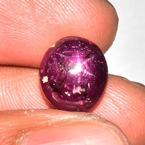 May include: A deep magenta-coloured ruby gemstone, oval in shape and polished to a high sheen. The stone displays a star effect across its surface. The gem is held between fingers, highlighting its colour and form.