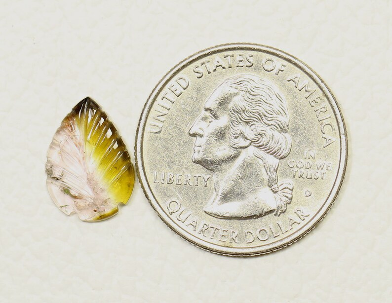 May include: A small, teardrop-shaped, carved gemstone with pink, yellow, and brown colors. The gemstone is next to a US quarter for size comparison.
