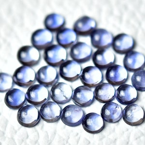 Könnte beinhalten: Eine Sammlung runder, glatter, violetter Edelsteine. Die Edelsteine sind lose auf einer weißen Oberfläche angeordnet.