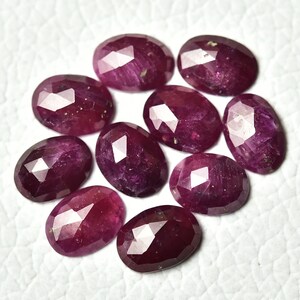 May include: Ten faceted oval ruby gemstones in various shades of pink and red. The gemstones are arranged in a cluster on a white surface.