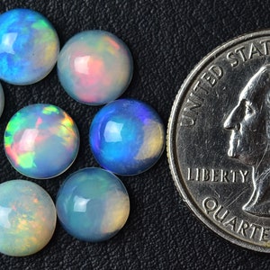 Pode incluir: Um grupo de oito caboch&otilde;es de opala iridescente, cada um com aproximadamente 1,25 cm de di&acirc;metro, dispostos em um c&iacute;rculo. Uma moeda de 25 centavos de d&oacute;lar americano est&aacute; inclu&iacute;da para compara&ccedil;&atilde;o de tamanho.