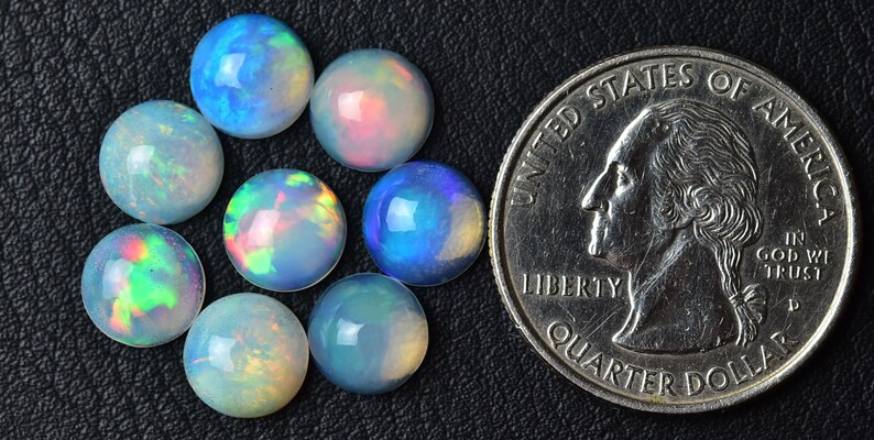 Pode incluir: Um grupo de oito caboch&otilde;es de opala iridescente, cada um com aproximadamente 1,25 cm de di&acirc;metro, dispostos em um c&iacute;rculo. Uma moeda de 25 centavos de d&oacute;lar americano est&aacute; inclu&iacute;da para compara&ccedil;&atilde;o de tamanho.