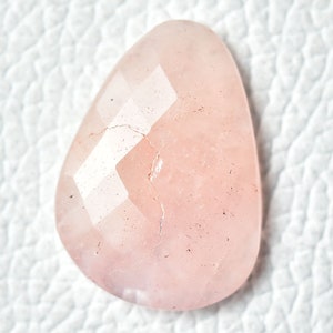 May include: A pale pink, faceted gemstone with a natural, irregular shape. The stone has a translucent appearance and shows signs of natural inclusions.