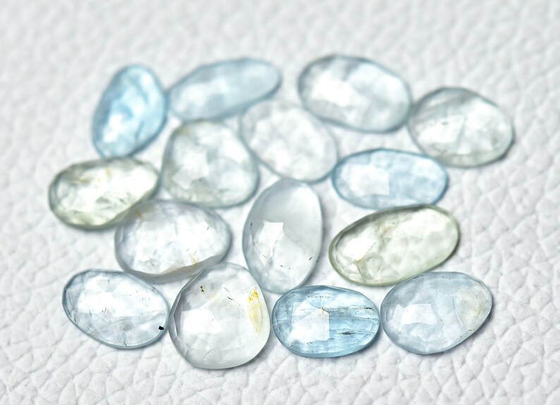 Puede incluir: Una colecci&oacute;n de piedras preciosas de aguamarina azul claro, cada una con una forma ovalada facetada. Las piedras est&aacute;n dispuestas sobre una superficie blanca.