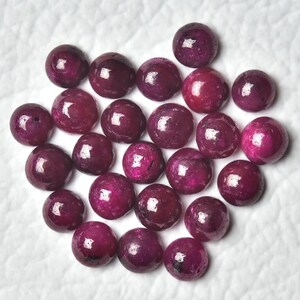May include: A collection of 25 round, cabochon cut ruby gemstones. The stones are a deep, rich red color and have a smooth, polished finish.