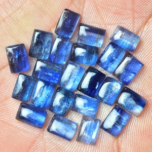 Può includere: Una collezione di 18 cabochon rettangolari di cianite blu. Le pietre sono traslucide e hanno un colore blu chiaro con alcune striature blu più scure.