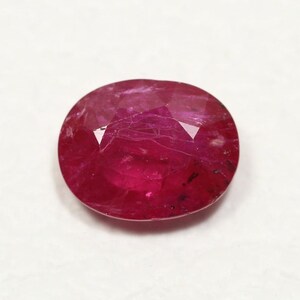 May include: A deep red oval-shaped gemstone with a faceted cut. The gemstone has a slight shimmer and is set against a white background.