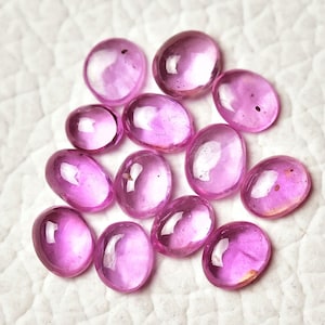 Può includere: Un gruppo di 12 pietre preziose rosa di forma ovale, probabilmente rubini, esposte su una superficie bianca.