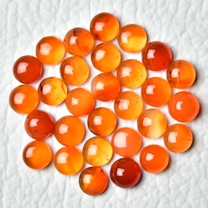 Pu&ograve; includere: Una collezione di gemme rotonde e lucide nei toni dell'arancione e dell'ambra. Le pietre sono disposte vicine, con variazioni di colore e traslucenza. Le gemme sono esposte su una superficie bianca e strutturata.