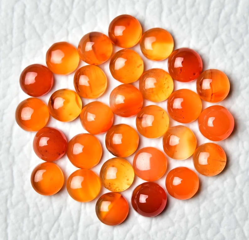 Pu&ograve; includere: Una collezione di gemme rotonde e lucide nei toni dell'arancione e dell'ambra. Le pietre sono disposte vicine, con variazioni di colore e traslucenza. Le gemme sono esposte su una superficie bianca e strutturata.