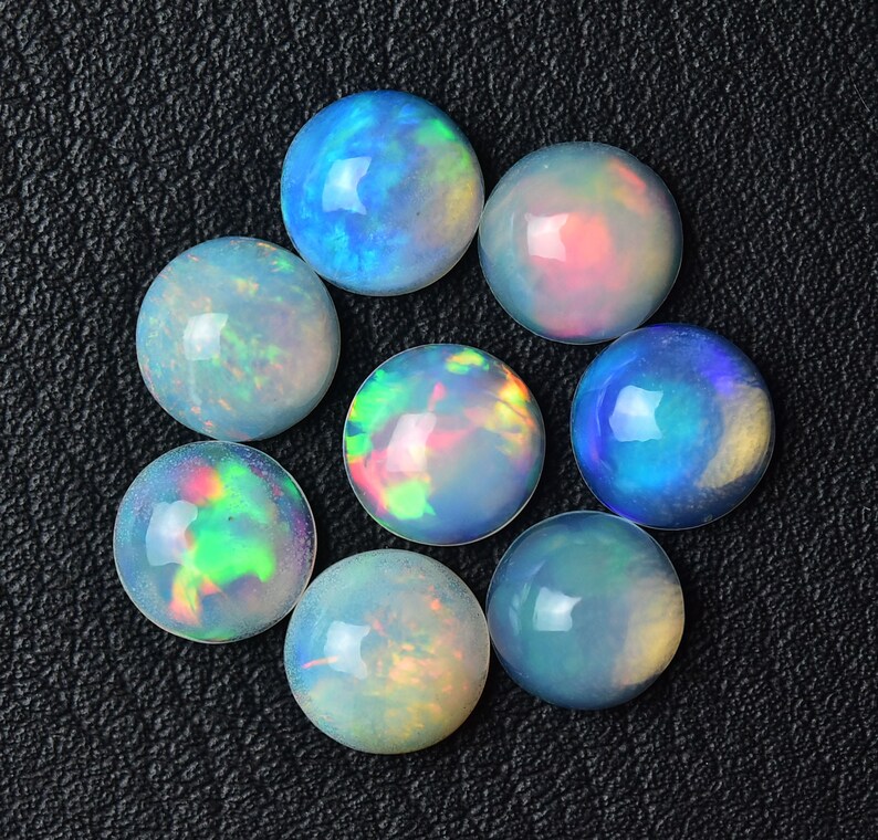 Pode incluir: Uma cole&ccedil;&atilde;o de oito caboch&otilde;es de opala redondos em v&aacute;rios tons de azul, verde e rosa. As opalas est&atilde;o colocadas sobre um fundo preto.