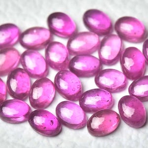 Pode incluir: Uma coleção de pedras preciosas ovais polidas em tons de rosa e magenta. As pedras preciosas estão dispostas juntas, criando um grupo de pedras vibrantes e translúcidas. As pedras são provavelmente rubis, para joalharia.