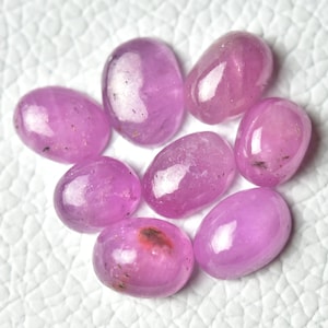 May include: Eight oval-shaped pink gemstones with a shiny, translucent finish. The gemstones are arranged in a cluster on a white surface.