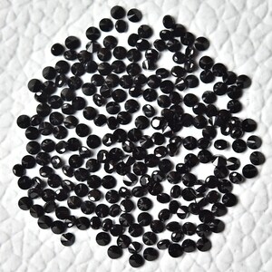 May include: A large pile of black round gemstones with faceted surfaces. The gemstones are scattered on a white surface.