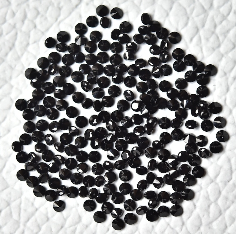 May include: A large pile of black round gemstones with faceted surfaces. The gemstones are scattered on a white surface.