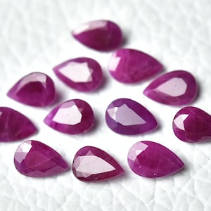 Peut inclure: Plusieurs rubis en forme de goutte, taillés à facettes. Les pierres précieuses sont d'un rouge-rose profond et sont disposées sur une surface blanche. Les facettes captent la lumière, créant des reflets.