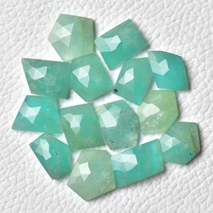 Puede incluir: Un conjunto de gemas facetadas de color verde azulado claro con diversas formas geométricas. Las gemas están dispersas sobre una superficie blanca, mostrando sus superficies pulidas y su claridad interna. La paleta de colores es suave y atractiva.